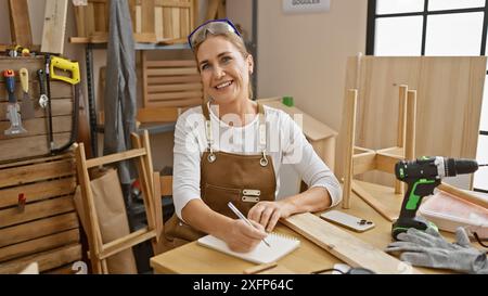 Una donna sorridente in un'officina di legno che scrive appunti con gli attrezzi intorno a lei e una sedia di legno in corso Foto Stock