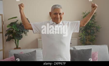 Uomo ispanico dai capelli grigi che flessa i muscoli in un salotto, mostrando sicurezza e vitalità in un ambiente domestico. Foto Stock