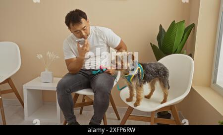 Un giovane asiatico siede in una sala d'attesa di una clinica veterinaria con il suo cane, conversando su un telefono cellulare mentre il suo cane sta su una sedia. Foto Stock