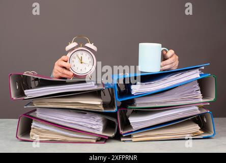 Donna stanca esausta con caffè e sveglia, una pila di cartelle di file, esaurimento, stress e sovraccarico, pressione sul lavoro Foto Stock