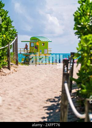 Luminoso vista panoramica di classic duna di sabbia il percorso foderato con vegetazione tropicale che conduce alla vibrante blu del mare con bagnino torre sulla spiaggia di South Beach a Miami Foto Stock