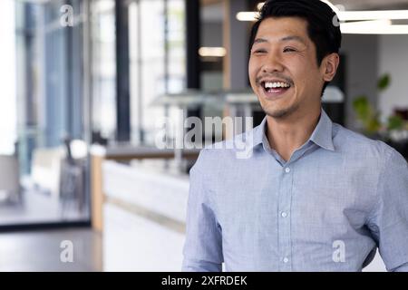 Uomo sorridente in ufficio che indossa una camicia blu, in piedi e con un aspetto felice, copia spazio Foto Stock