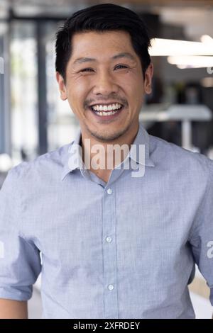 Un uomo sorridente in ufficio che indossa una camicia blu, guarda la macchina fotografica e si sente felice Foto Stock