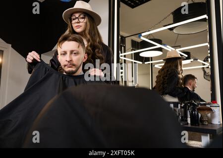 Un giovane con la barba in un mantello nero siede su una sedia in un barbiere. Il cliente sta aspettando un taglio di capelli e la rasatura della barba. Foto Stock