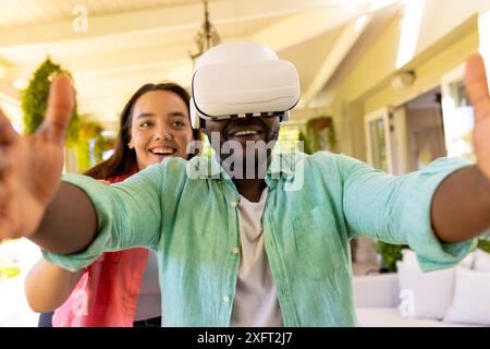 Utilizzando il visore VR, l'uomo si gode un'esperienza di realtà virtuale con un amico sorridente nelle vicinanze Foto Stock