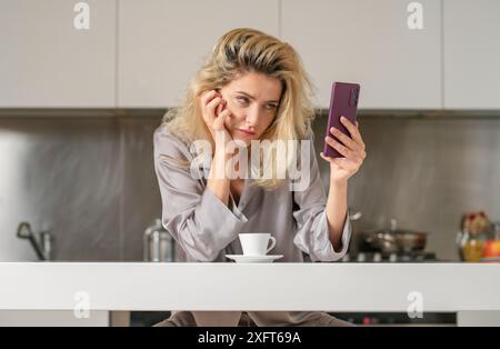 Ragazza sconvolta con il telefono bere vino in cucina. Mattina di bella donna. Signora triste che usa lo smartphone. Concetto di social media. Foto Stock