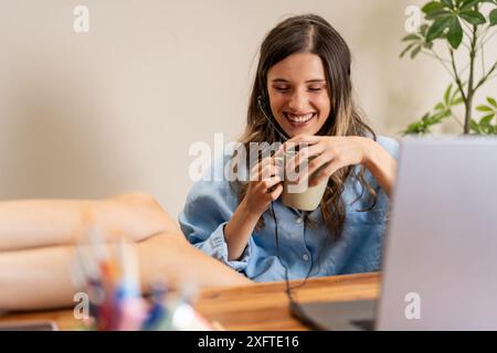 Una donna sorridente con una cuffia che si rilassa nel suo ufficio di casa, si alza sulla scrivania e si gode una pausa caffè. Configurazione del lavoro in remoto comoda e informale Foto Stock