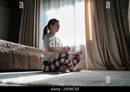 Una donna siede a gambe incrociate sul pavimento, meditando in una stanza luminosa con la luce del sole che scorre attraverso la finestra, creando un'atmosfera tranquilla. Foto Stock