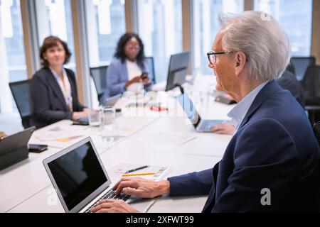 Uomo d'affari anziano che utilizza un computer portatile mentre ascolta i colleghi durante una riunione in ufficio Foto Stock