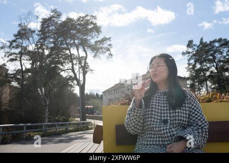 Donna che fuma penna svapo mentre è seduta sulla panchina durante la giornata di sole Foto Stock