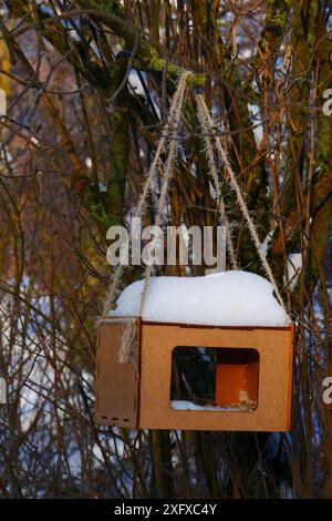 Un alimentatore di legno ricoperto di neve su una fune è appeso ai rami di un albero. Foto Stock