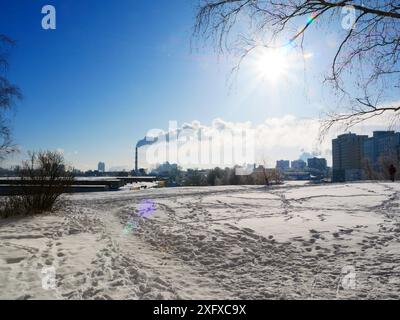 Un campo ricoperto di neve con molte impronte, all'orizzonte di un camino fumante sullo sfondo di un cielo azzurro e soleggiato. Concetto ecologico. Foto Stock