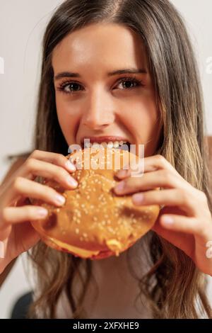 Donna che mangia hamburger - gusta fast food - pasto delizioso, donna sorridente - indulgenza nel cibo spazzatura, cena informale - gustoso hamburger. Foto Stock