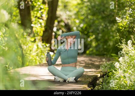 donna sportiva che tiene in mano una bottiglia d'acqua e si pulisce la fronte dopo essersi allenata all'aperto in un parco Foto Stock