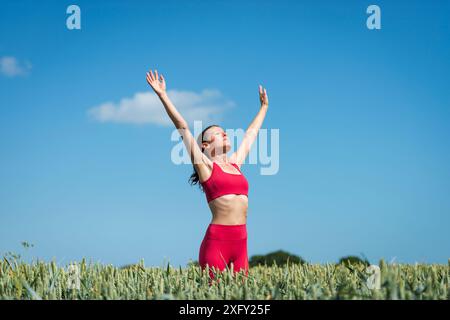 Donna sportiva con le braccia alzate per festeggiare e godersi il sole nel campo del grano, indossando abbigliamento sportivo rosso con il cielo blu. Foto Stock