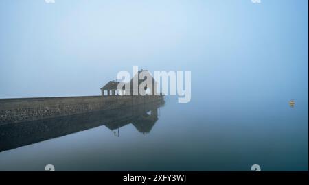 Parete della diga sul lago Edersee nella nebbia mattutina, riflesso nelle acque calme, una boa gialla sul bordo della foto. Distretto di Waldeck-Frankenberg, Assia, Germania. Foto Stock