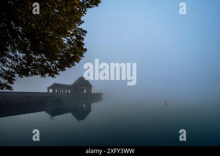 Parete della diga sul lago Edersee nella nebbia mattutina, riflesso nelle acque calme, una boa gialla sul bordo della foto. Distretto di Waldeck-Frankenberg, Assia, Germania. Foto Stock