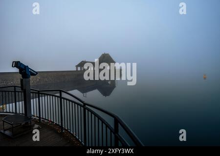 Parete della diga sul lago Edersee nella nebbia mattutina, riflesso nelle acque calme, una boa gialla sul bordo della foto. Visualizzazione del binocolo in primo piano. Distretto di Waldeck-Frankenberg, Assia, Germania. Foto Stock