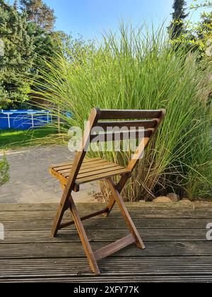 La sedia pieghevole in legno si erge sulla terrazza di fronte ad un cespuglio con erba verde di pampas in un giorno d'estate e sullo sfondo si può vedere una piscina Foto Stock