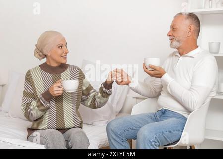 Donna matura dopo la chemioterapia e suo marito con tazze di tè che tengono per mano in camera da letto Foto Stock