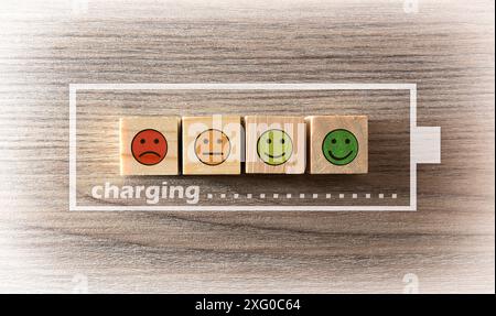 Concetto di ricarica di energia vitale con blocchi di legno con emoticon con espressioni su tavolo in legno, simboli della batteria e messaggio di ricarica. Foto Stock