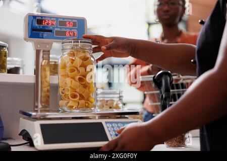 Primo piano del fornitore afroamericano che utilizza una bilancia digitale per misurare il peso del prodotto organico fresco in un contenitore di vetro. Vista dettagliata del vasetto di pesatura commerciante di pasta biologica mentre il cliente attende al bancone. Foto Stock