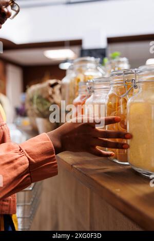 Donna afroamericana che acquista verdure fresche e biologiche in un negozio di alimentari. L'immagine ravvicinata mostra la mano di una signora nera su contenitori di vetro con pasta. Opzioni sostenibili, confezione riutilizzabile. Foto Stock