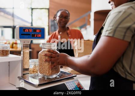 Una donna afroamericana in grembiule assiste un cliente, pesando grani di provenienza locale su scala presso un negozio di alimentari fresco e biologico. Vaschetta di misurazione per commercianti locali riempita con prodotti alimentari biologici. Foto Stock