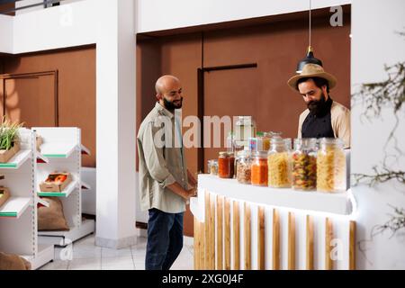 Un uomo del Medio Oriente si trova al banco cassa. Questo bancone è pieno di vari alimenti in confezioni ecologiche. Dall'altra parte c'è un lavoratore di negozio maschile caucasico che indossa un cappello. Foto Stock