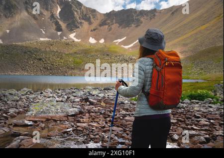 Vista posteriore dell'elegante hipster donna con zaino e cappello grigio che guarda al lago di montagna durante le attività ricreative all'aperto. Rilassatevi mentre vi rilassate su un concetto Foto Stock
