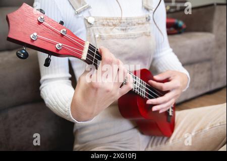 Primo piano, ukulele in mani femminili, hipster impara a suonare uno strumento musicale a casa, seduto sul pavimento vicino al divano Foto Stock