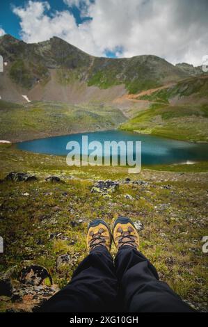 Piedi di un turista alpino in scarpe sullo sfondo di verdi montagne e di un lago azzurro. escursioni, solitudine Foto Stock