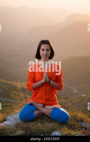 Yogini in posizione di loto in pieno volto siede su una scogliera sullo sfondo del cielo del tramonto con montagne, prega Foto Stock