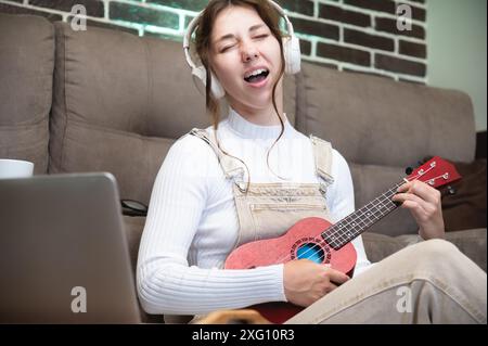 Giovane studentessa che impara la voce e suona l'ukulele a casa seduta sul pavimento nel soggiorno di fronte a un laptop Foto Stock