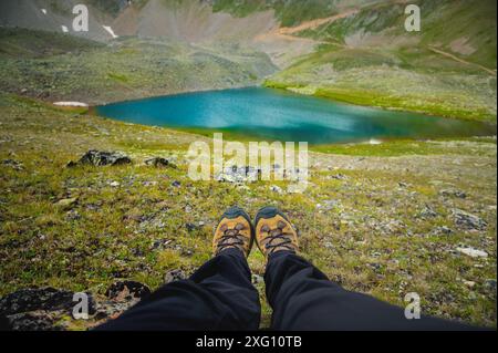 Il viaggiatore riposa su un altopiano di montagna. Vista in prima persona, gambe ravvicinate sullo sfondo di un lago turchese di montagna Foto Stock