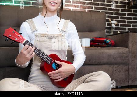 Primo piano di mani femminili che suonano l'ukulele rosso seduto a casa sullo sfondo del divano Foto Stock