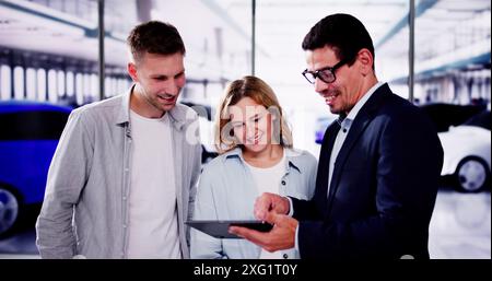 Giovane coppia sorridente mentre si trova di fronte a una nuova auto presso la concessionaria, tenendo la chiave e firmando un contratto con il venditore. Foto Stock