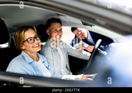 Il venditore di auto professionale espone con entusiasmo un nuovo veicolo elegante alla concessionaria interna per giovani coppie, donna ispanica che tiene il volante sorridente. Foto Stock