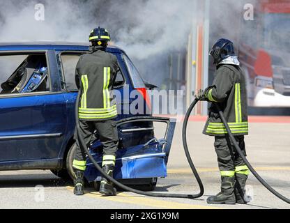 i vigili del fuoco in uniforme con caschi estinguono l'incendio dell'auto distrutta dopo l'incidente con il grande tubo flessibile sulla strada Foto Stock