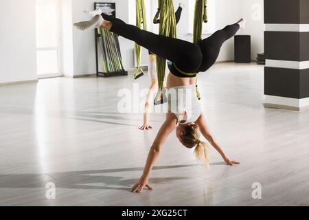 una donna esegue una posa invertita impegnativa durante una lezione di yoga aereo in una palestra luminosa e moderna. Foto Stock