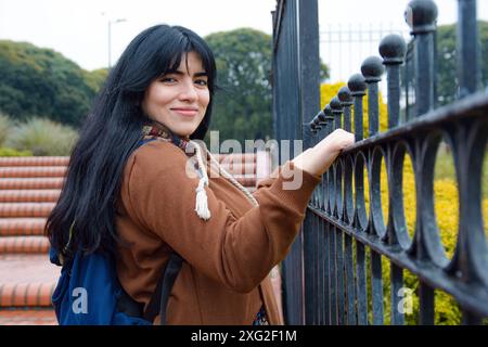 vista laterale della giovane donna latina che tiene la recinzione del parco guardando la telecamera Foto Stock