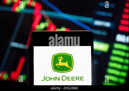 In questa illustrazione fotografica, la società americana che produce macchinari agricoli, attrezzature pesanti e macchinari forestali, John Deere e nota anche come Deere & Company (NYSE: DE), logo visualizzato su uno smartphone con un grafico indice di borsa economica sullo sfondo. Foto Stock