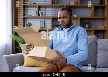 Un uomo è seduto su un divano grigio in un salotto moderno, sembra scioccato mentre apre una scatola di cartone. Lo sfondo presenta una libreria in legno con libri e oggetti decorativi. Foto Stock