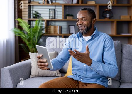 Uomo sorridente che indossa abiti casual e che fa una chiacchierata amichevole sul tablet mentre è seduto sul divano in un salotto accogliente. L'immagine trasmette concetti di comunicazione, tecnologia. Foto Stock