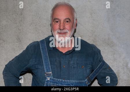 Un senior con tute in denim e una camicia blu si erge casualmente davanti a una parete di cemento, sorridendo davanti alla fotocamera. Foto Stock