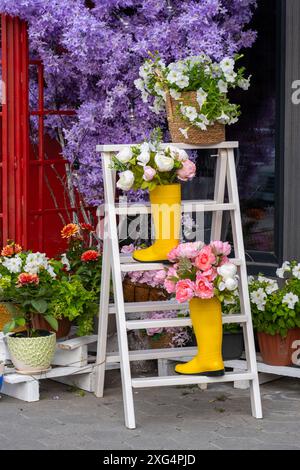 Bouquet di fiori in stivali di gomma gialli su piedistallo in legno. Negozio di fiori con facciata splendidamente arredata Foto Stock