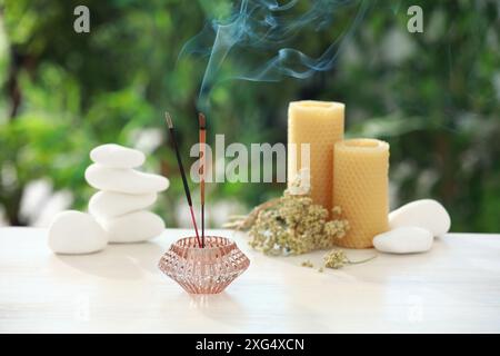 Bastoncini di incenso che si mescolano nel supporto vicino a pietre, candele e fiori secchi su un tavolo di legno all'aperto Foto Stock