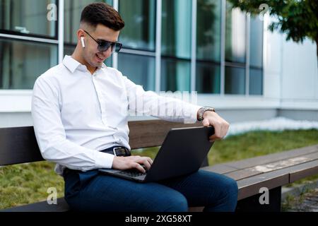 Un uomo è seduto su una panchina all'aperto, concentrato sullo schermo del suo computer portatile. Sembra impegnato nella digitazione o nella navigazione. Foto Stock