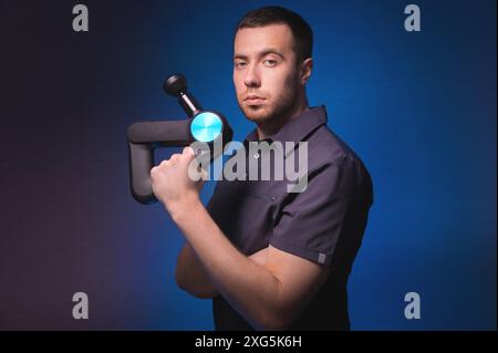 Ritratto di un massaggiatore professionista sicuro di sé con un massaggiatore a percussione in mano. Tasto basso, sfondo blu. Massaggio con onde d'urto Foto Stock