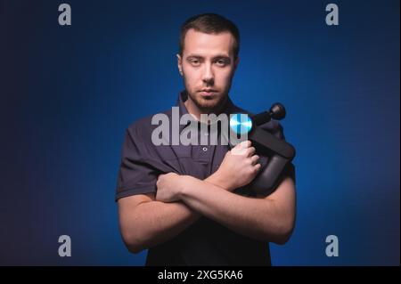 Ritratto di un massaggiatore professionista sicuro di sé con un massaggiatore a percussione in mano. Tasto basso, sfondo blu. Massaggio con onde d'urto Foto Stock